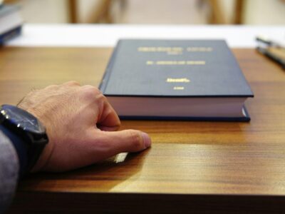Close-up of a left hand with a black wristwatch resting on a wooden desk beside a thick blue hardcover book.