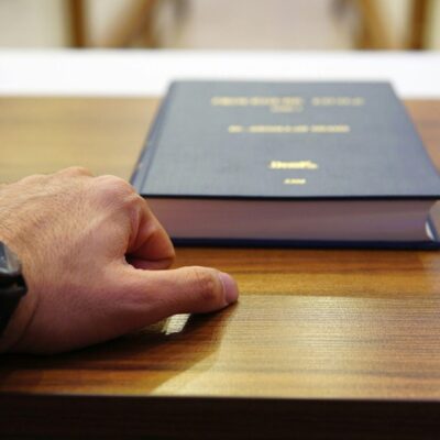 Close-up of a left hand with a black wristwatch resting on a wooden desk beside a thick blue hardcover book.