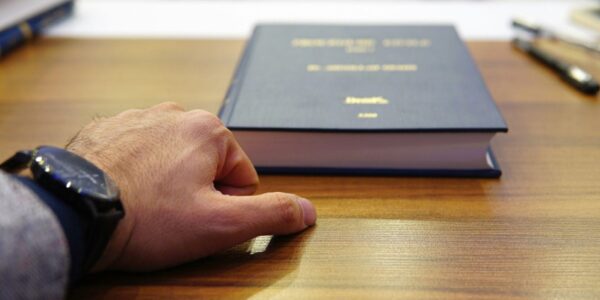 Close-up of a left hand with a black wristwatch resting on a wooden desk beside a thick blue hardcover book.
