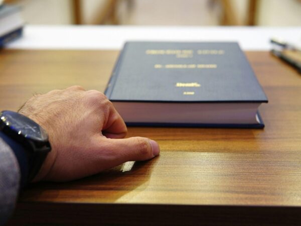 Close-up of a left hand with a black wristwatch resting on a wooden desk beside a thick blue hardcover book.